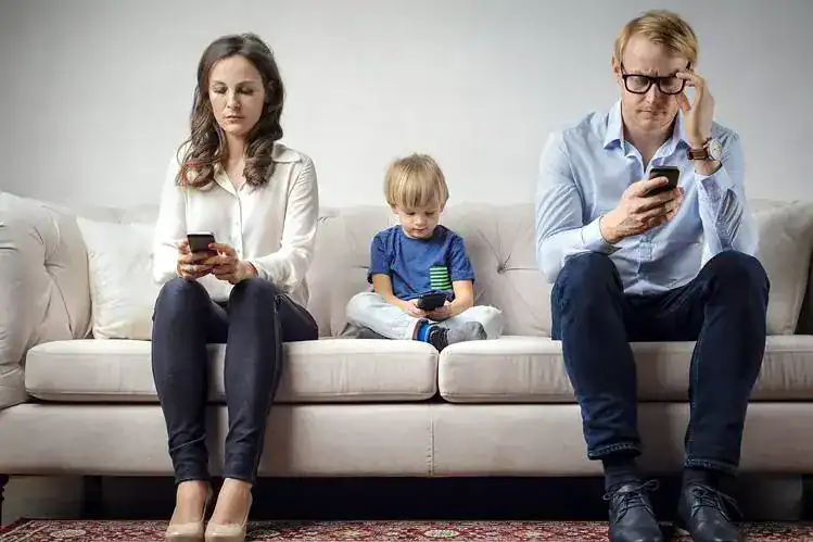 Family sitting on sofa, each using a smartphone, with a young boy in the middle.