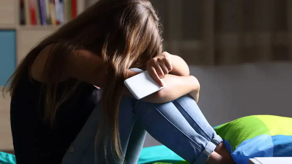 Person sitting on bed with head down, hugging knees, in a dimly lit room.