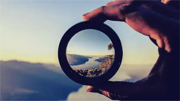 Person holding a circular lens showing a scenic mountain view at sunset.
