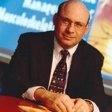 Bald man in glasses wearing a suit and colorful tie, sitting at a desk.